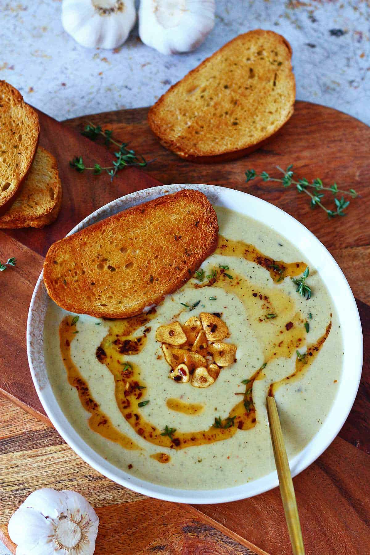 Roasted garlic soup in a bowl with toasted bread and thyme on a wooden board.