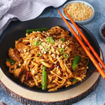 A black shallow bowl of kung pao chicken noodles with chopsticks on a wood slab.