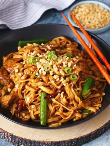 A black shallow bowl of kung pao chicken noodles with chopsticks on a wood slab.