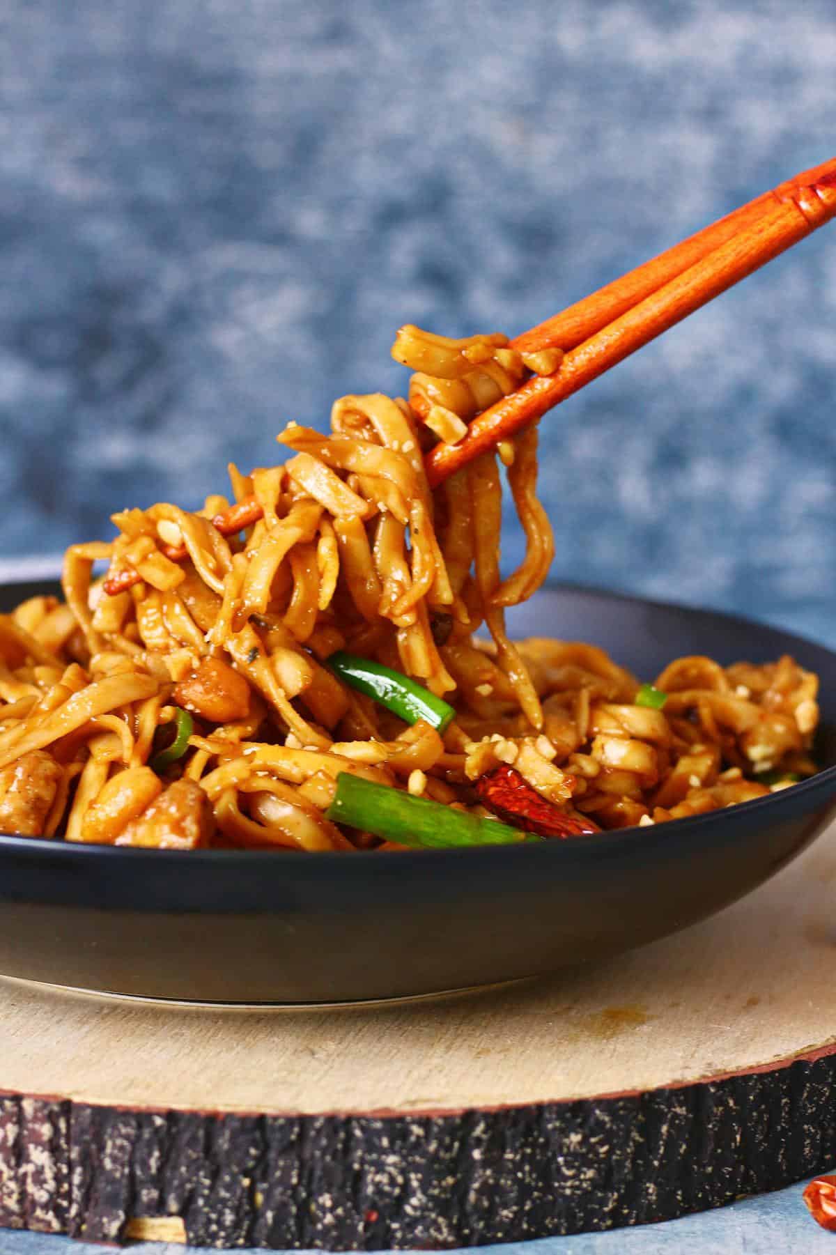 Picking up kung pao noodles with chopsticks from a black shallow plate.