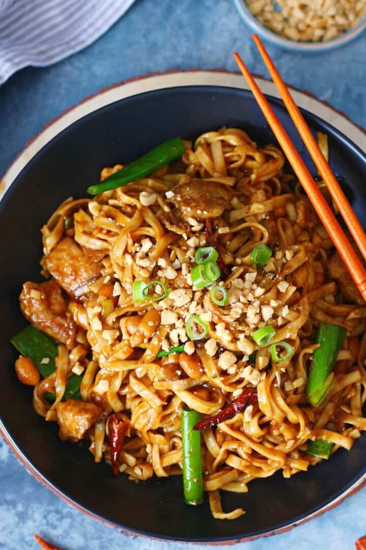 Overhead view of Kung Pao chicken noodles in a shallow black plate with chopsticks, with chopped peanuts in a small bowl in the background.
