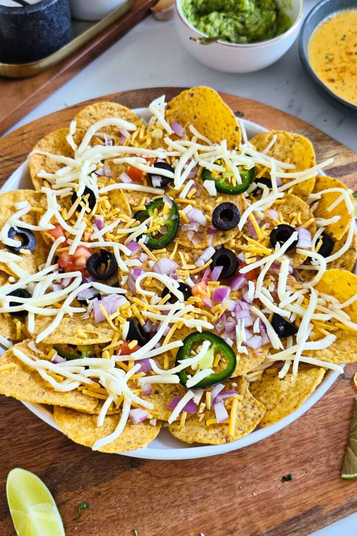 Layered veggie nachos with shredded cheddar and mozzarella, olives, jalapeรฑos, and red onion in a baking dish before baking.