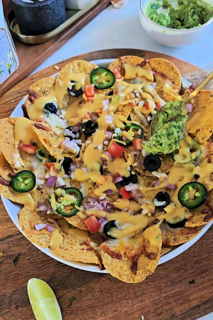 Fresh guacamole being spooned over baked cheesy veggie nachos with jalapeรฑos and olives.