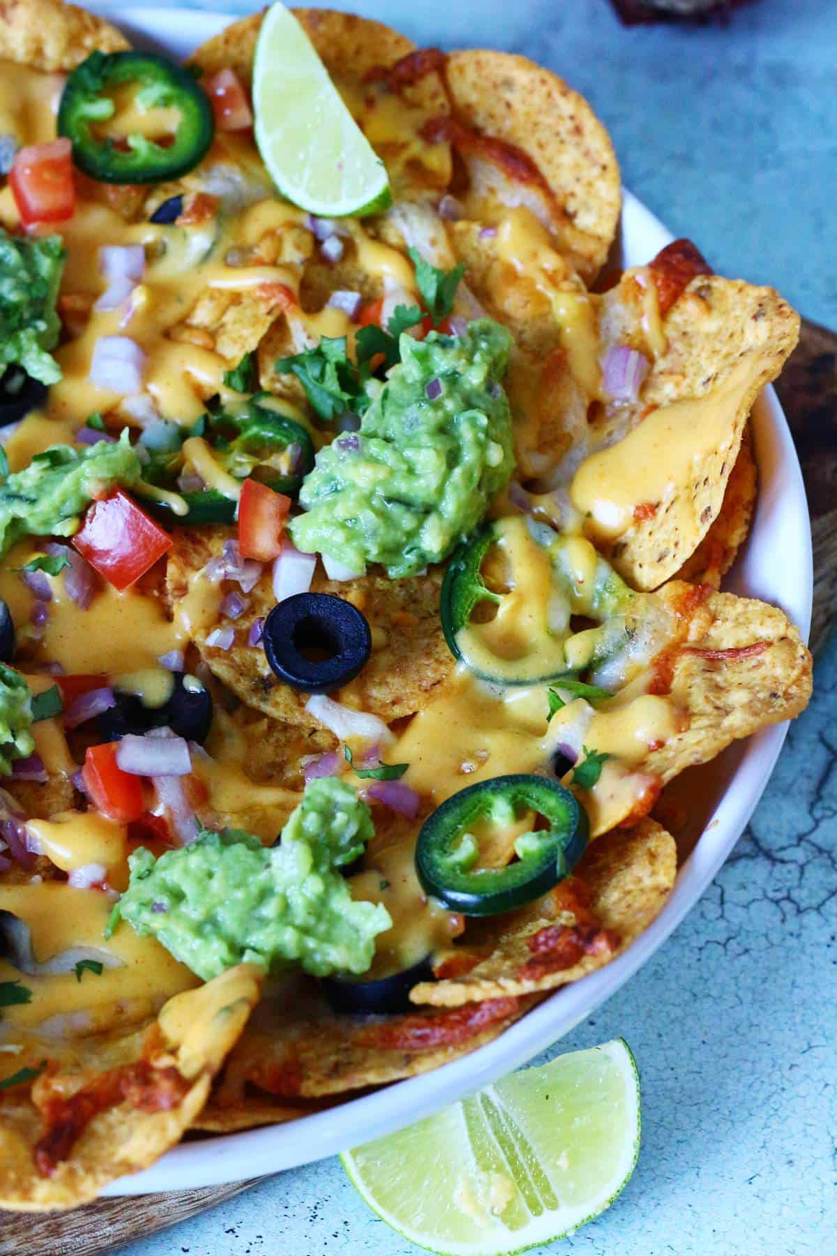 Close-up of cheesy veggie nachos with guacamole, jalapeรฑo slices, red onion, and olives.