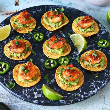 Mini shrimp tostadas on a black serving plate topped with guacamole, shrimp and cilantro.