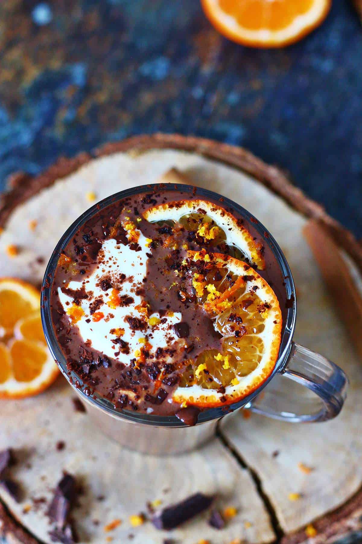 Close-up of orange hot chocolate topped with whipped cream, orange zest, and chocolate shavings, with sliced oranges in the background.