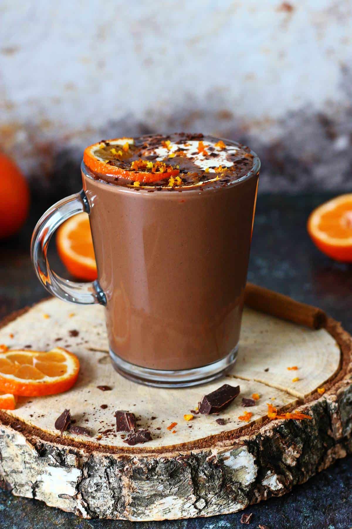 A mug of creamy orange hot chocolate on a wooden slab, garnished with orange slices and chocolate pieces.