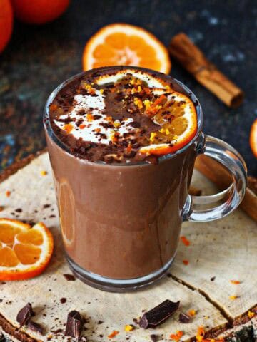Orange hot chocolate served in a clear mug, topped with orange slices, whipped cream, and chocolate shavings, surrounded by fresh oranges and cinnamon.