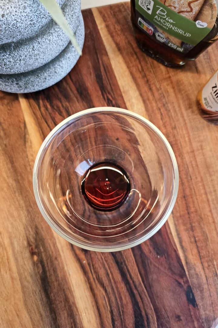 Maple syrup added to the bottom of a clear glass on a wooden surface.