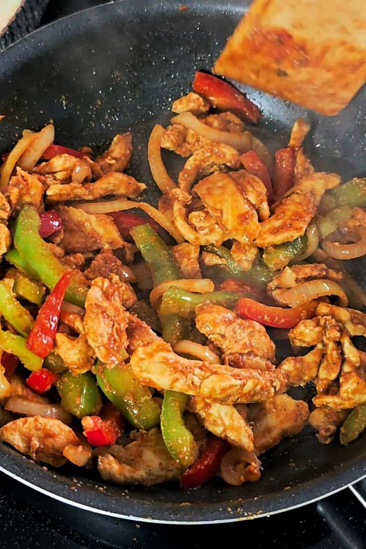 Chicken fajita filling with onions and bell peppers cooking in a black pan.