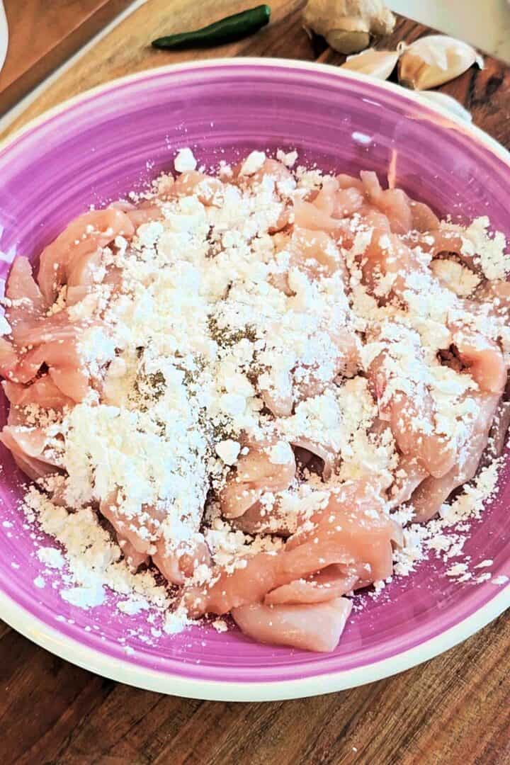 Raw chicken strips in a bowl mixed with cornflour, black pepper, and salt.
