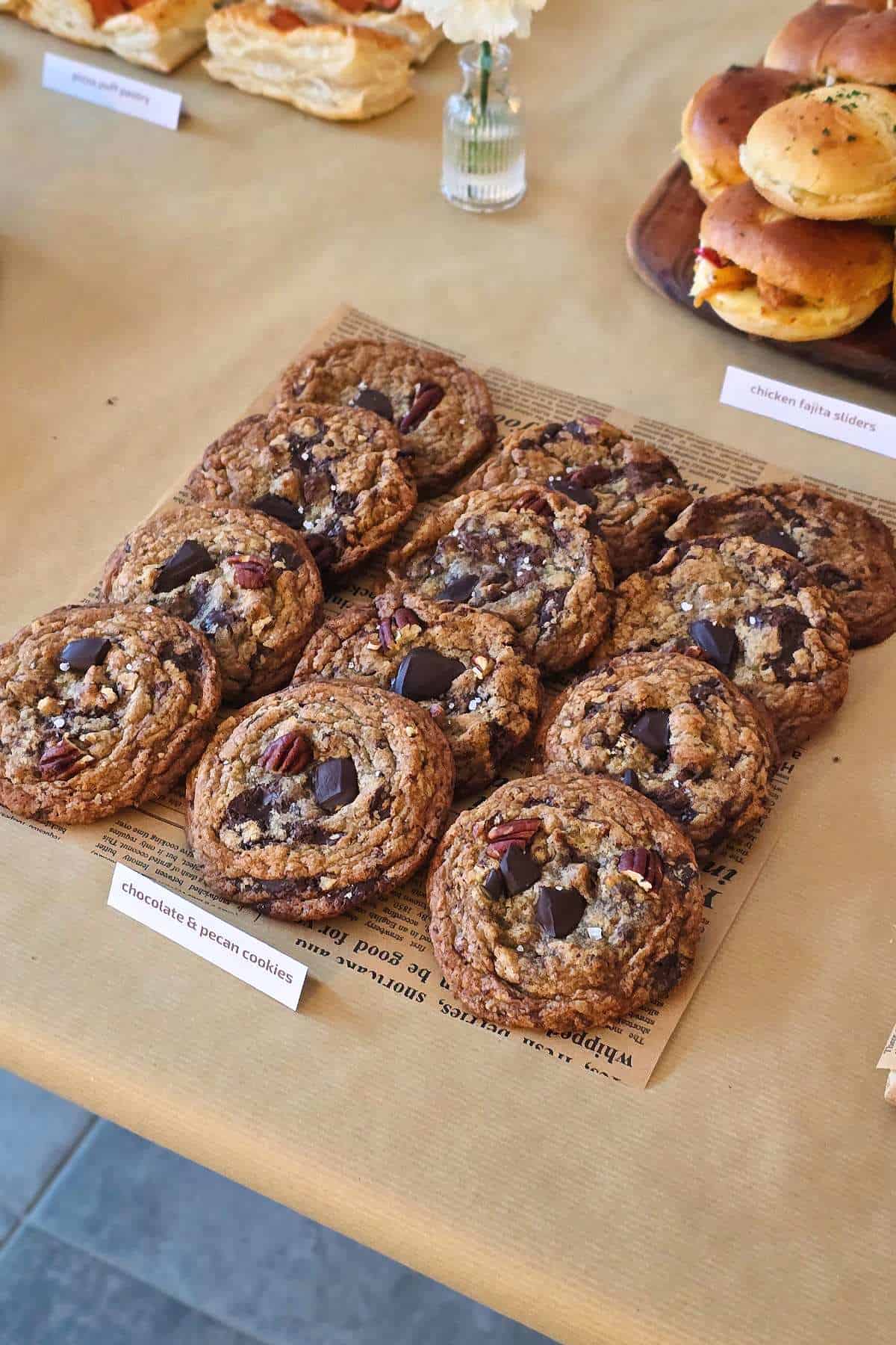 Chocolate chip pecan cookies arranged on parchment paper at a café-style table with sliders and pastries in the background.