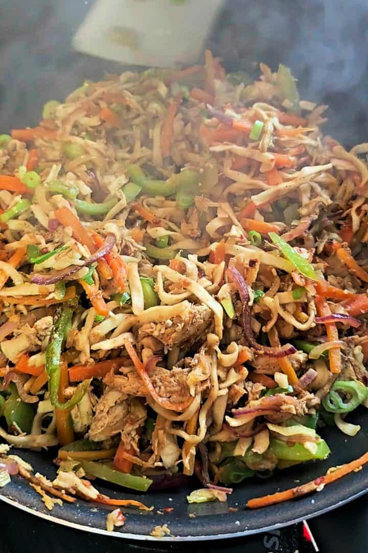 Chicken, noodles, carrots, bell peppers, and red onion stir-frying in a pan.