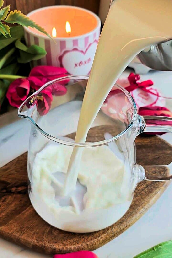Milk being poured into a clear glass jug placed on a wooden board.