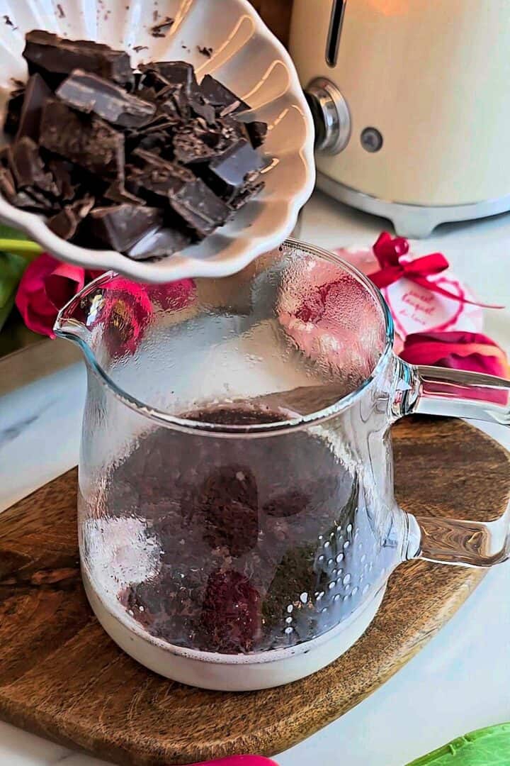 Chopped chocolate and cocoa powder being added to warm milk in a glass jug.