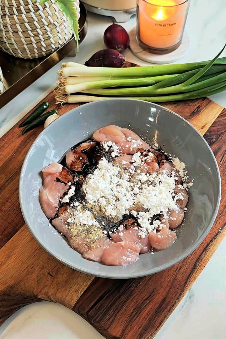 Raw chicken pieces in a bowl with soy sauce and cornstarch on top, placed on a wooden board with spring onions, garlic, and chilies nearby.