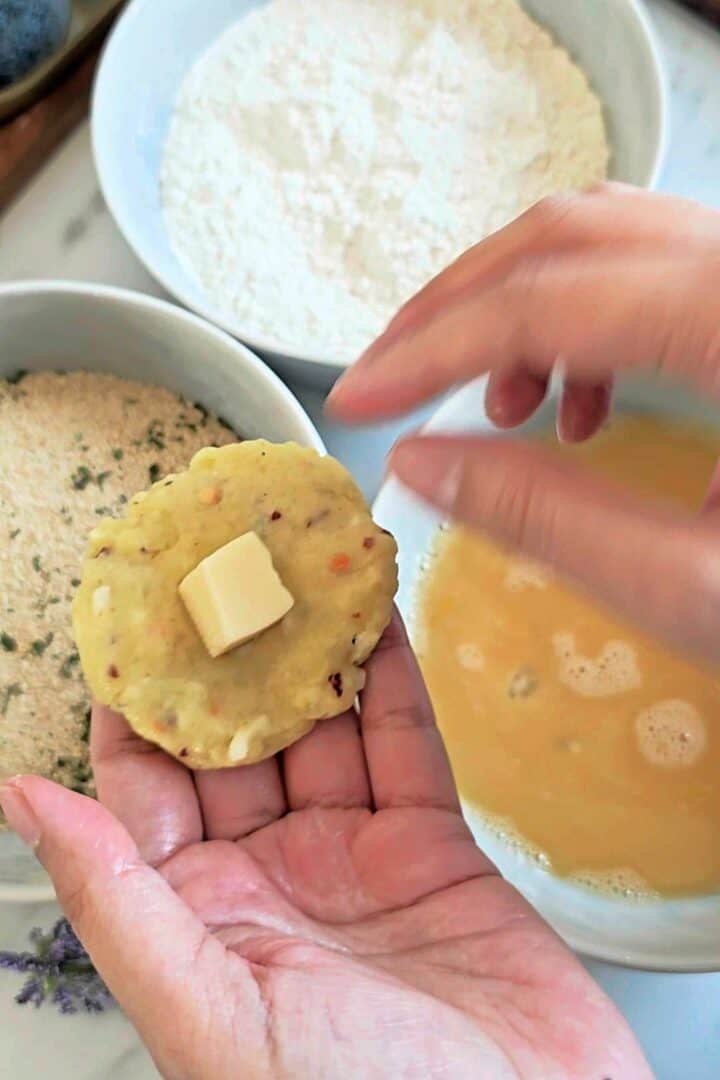 Hand holding flattened potato mixture with a cheese cube in the center next to bowls of flour, egg wash, and breadcrumbs.