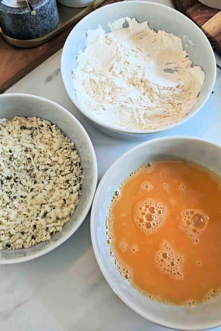 Three bowls containing flour, beaten egg, and breadcrumbs arranged on a counter.