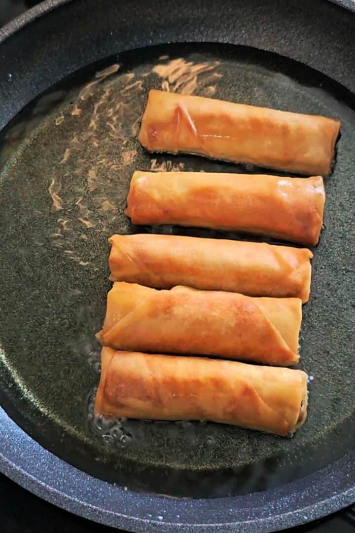 Spring rolls frying in a shallow pan of oil, turning golden brown.