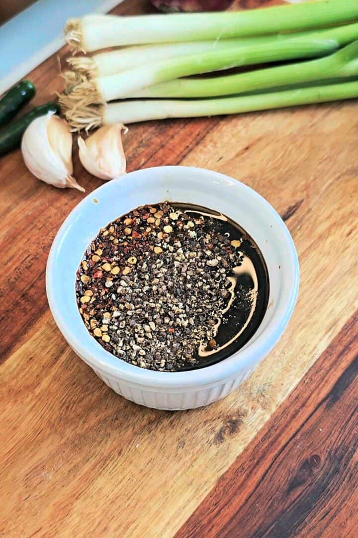 Small bowl of dark sauce topped with crushed black pepper and red chili flakes on a wooden board with garlic cloves and green chilies.