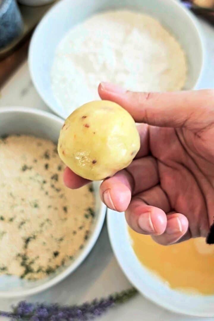 Hand holding a smooth, sealed potato ball before coating, with flour and egg wash bowls in the background.