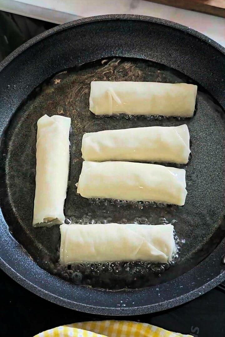 Uncooked spring rolls frying in shallow oil in a nonstick pan.