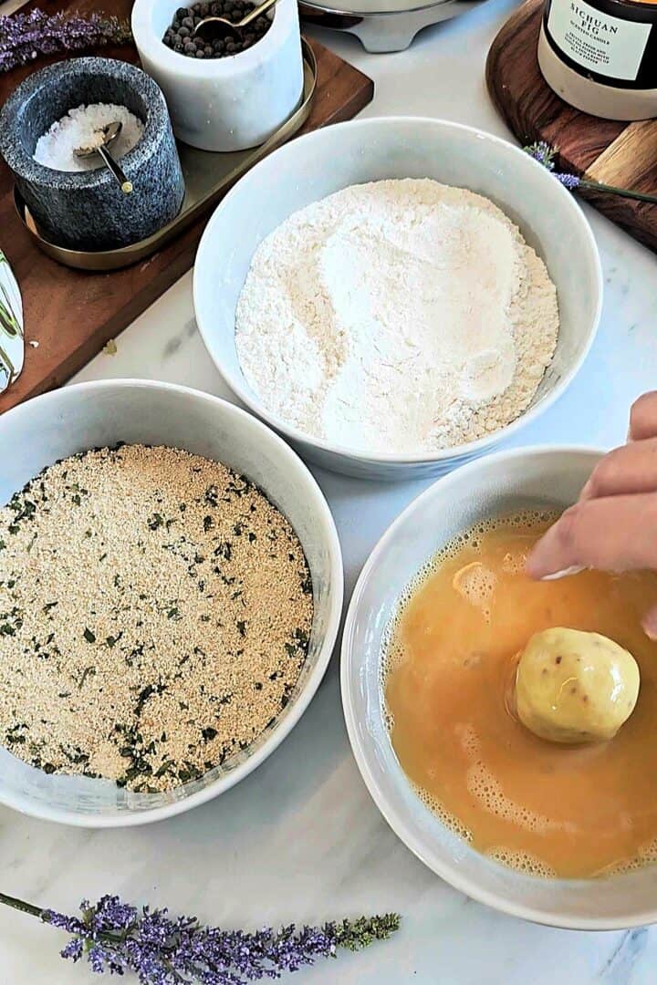 Three bowls arranged for coating with flour, seasoned breadcrumbs, and beaten egg, with a potato ball being dipped into egg wash.