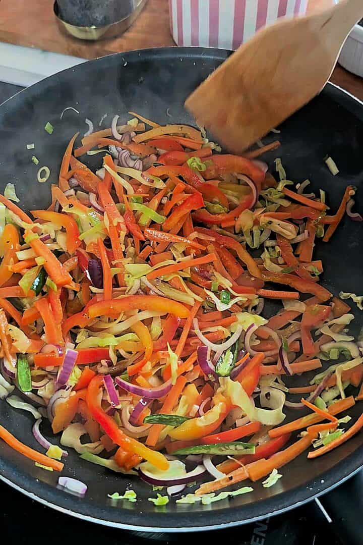 Sliced carrots, bell peppers, cabbage, and red onion being stir-fried in a pan with a wooden spatula.