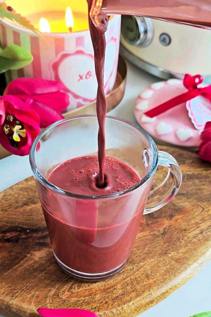 Red velvet hot chocolate being poured from a glass jug into a clear glass mug.