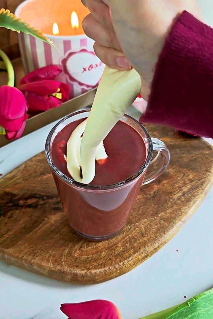 Cream cheese topping being piped onto red velvet hot chocolate in a glass mug.