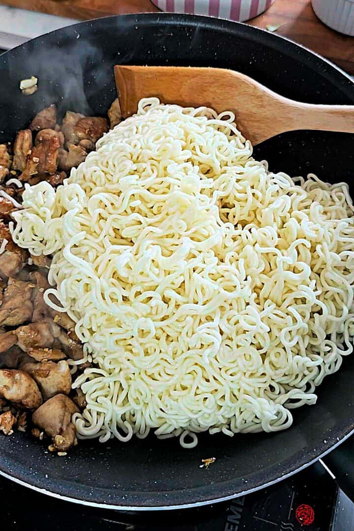 Cooked noodles added to a pan with browned chicken, with a wooden spatula resting on top.