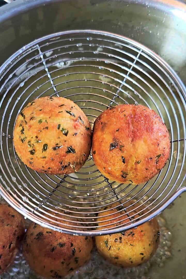 Two golden potato balls lifted from hot oil in a wire skimmer over a deep pot while frying.