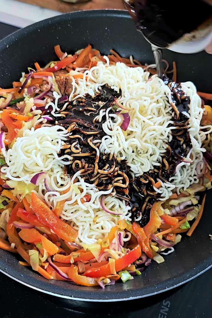 Dark chow mein sauce being poured over noodles and vegetables in a pan.