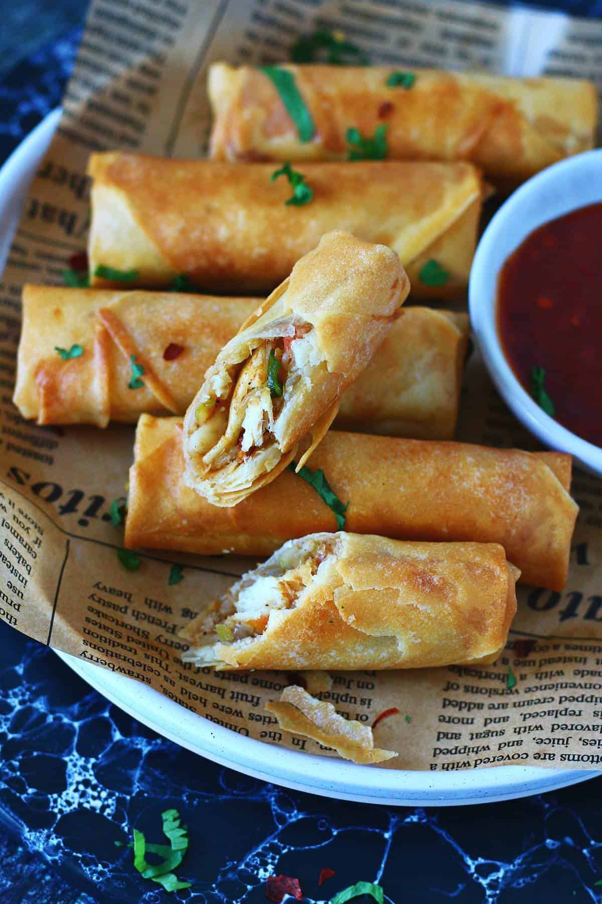 Spring rolls on a white plate lined with parchment paper, one roll cut open showing shredded chicken and vegetables, served with red dipping sauce.