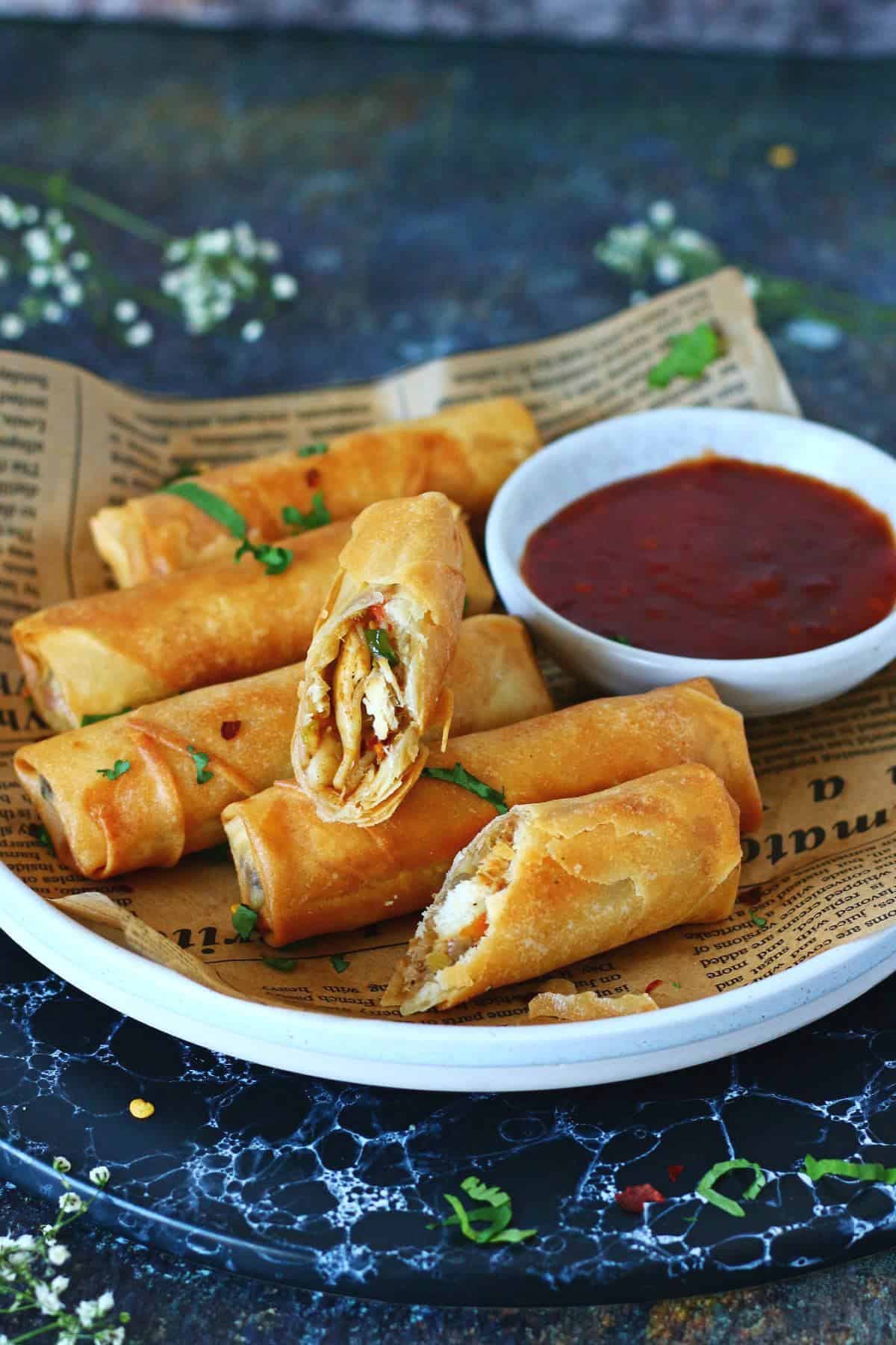 Plate of crispy chicken noodle spring rolls on parchment paper with a small bowl of red dipping sauce on the side.