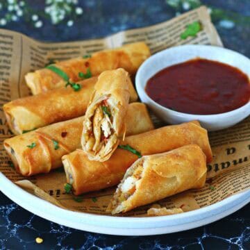 Crispy spring rolls stacked on a white plate, one roll sliced open to show chicken and noodle filling, with red sauce in a bowl.