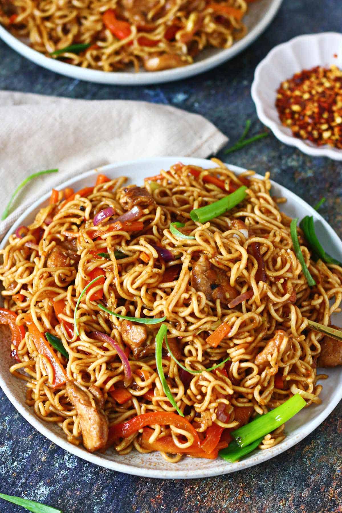 Chicken and vegetable chow mein in an ivory plate with red pepper flakes in the background.
