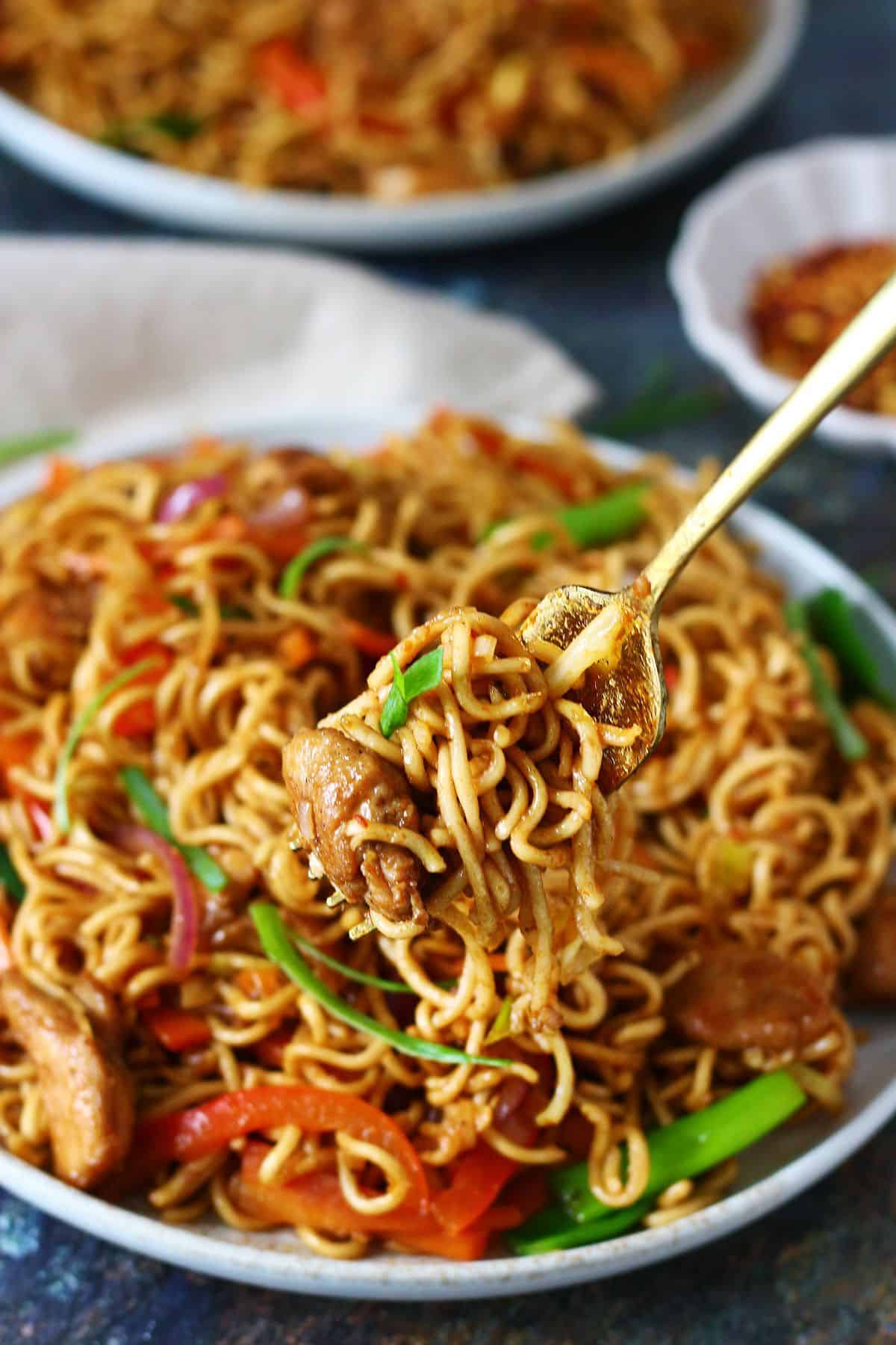 Fork lifting chicken chow mein with a small bowl of chili flakes in the background.