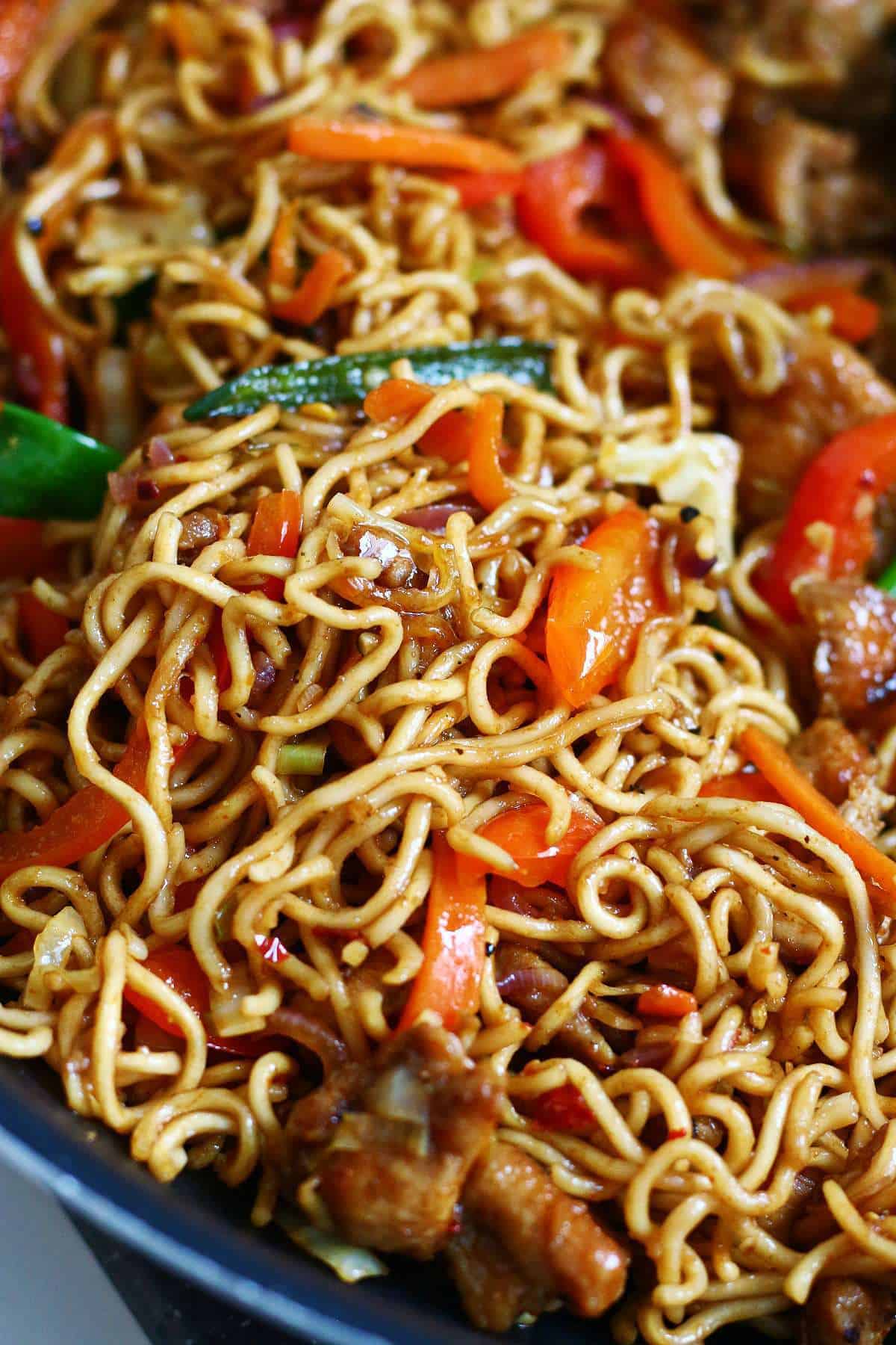 Close-up of chicken chow mein in a wok with glossy noodles, carrots, bell peppers, cabbage, and chicken pieces.