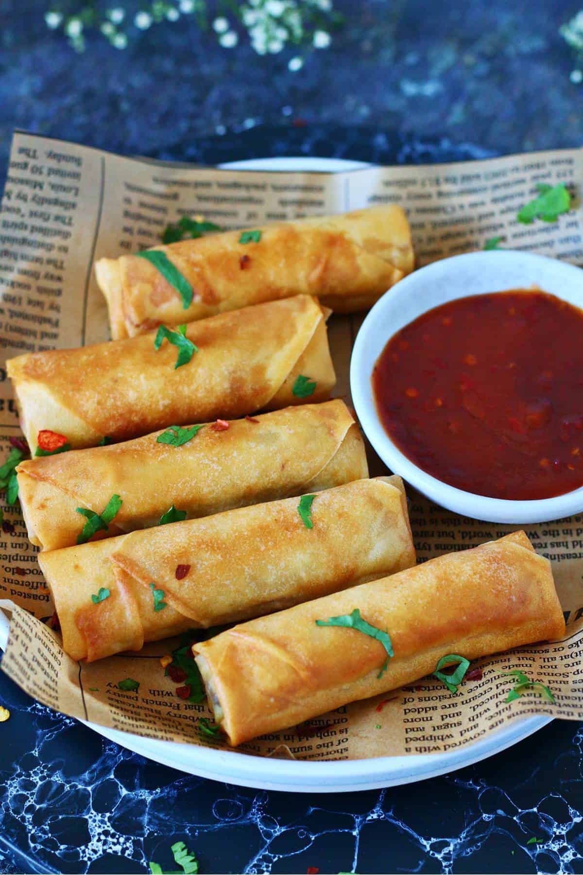 Five golden brown spring rolls arranged on parchment paper on a white plate with a bowl of red dipping sauce.
