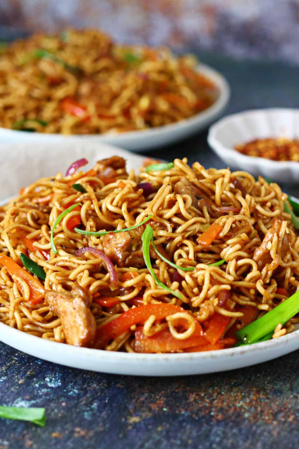 Plate of desi chicken chow mein garnished with spring onions, with another plate blurred in the background.