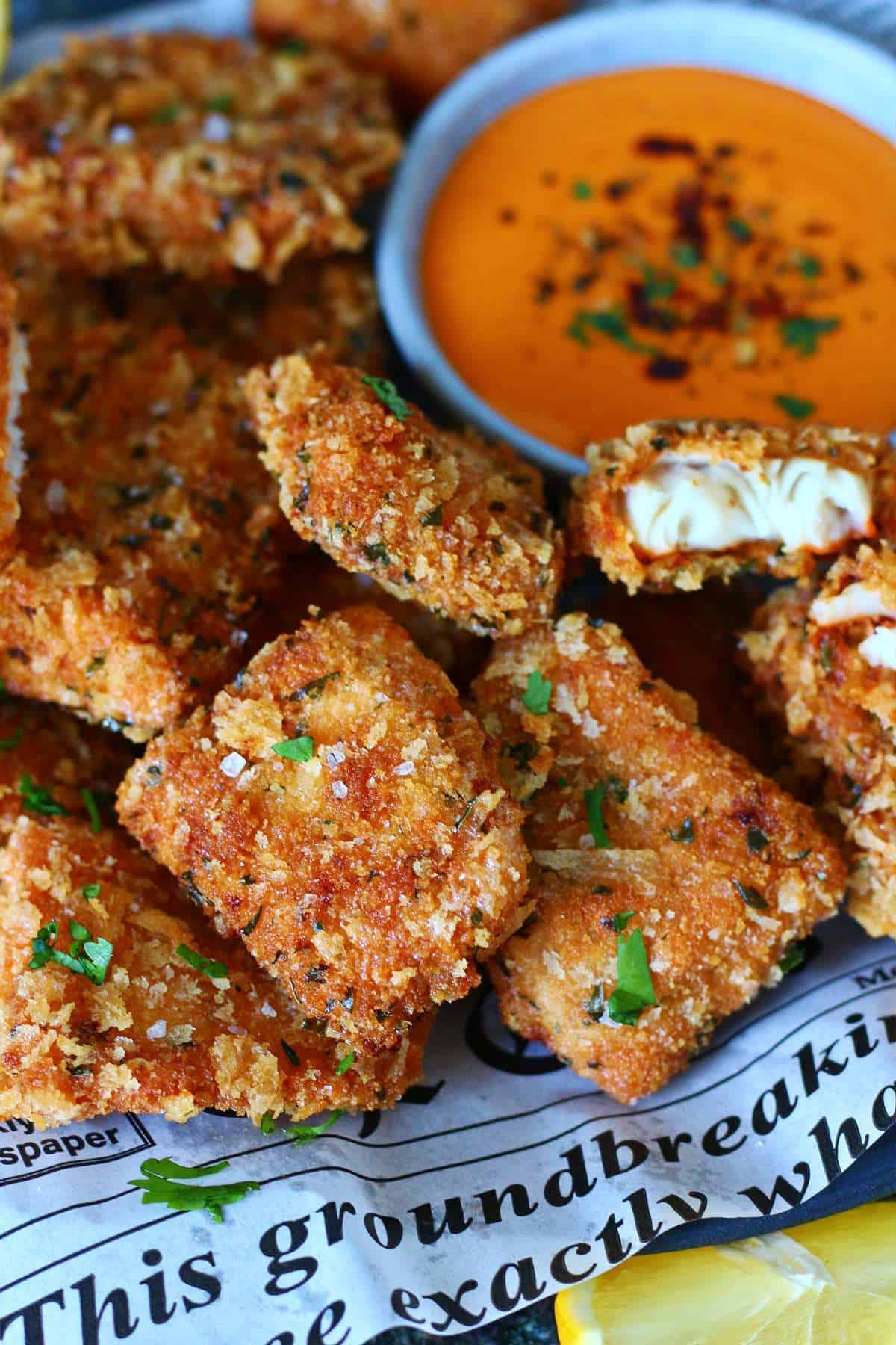 A broken breaded fish nugget showing the white fish inside next to a bowl of sauce.