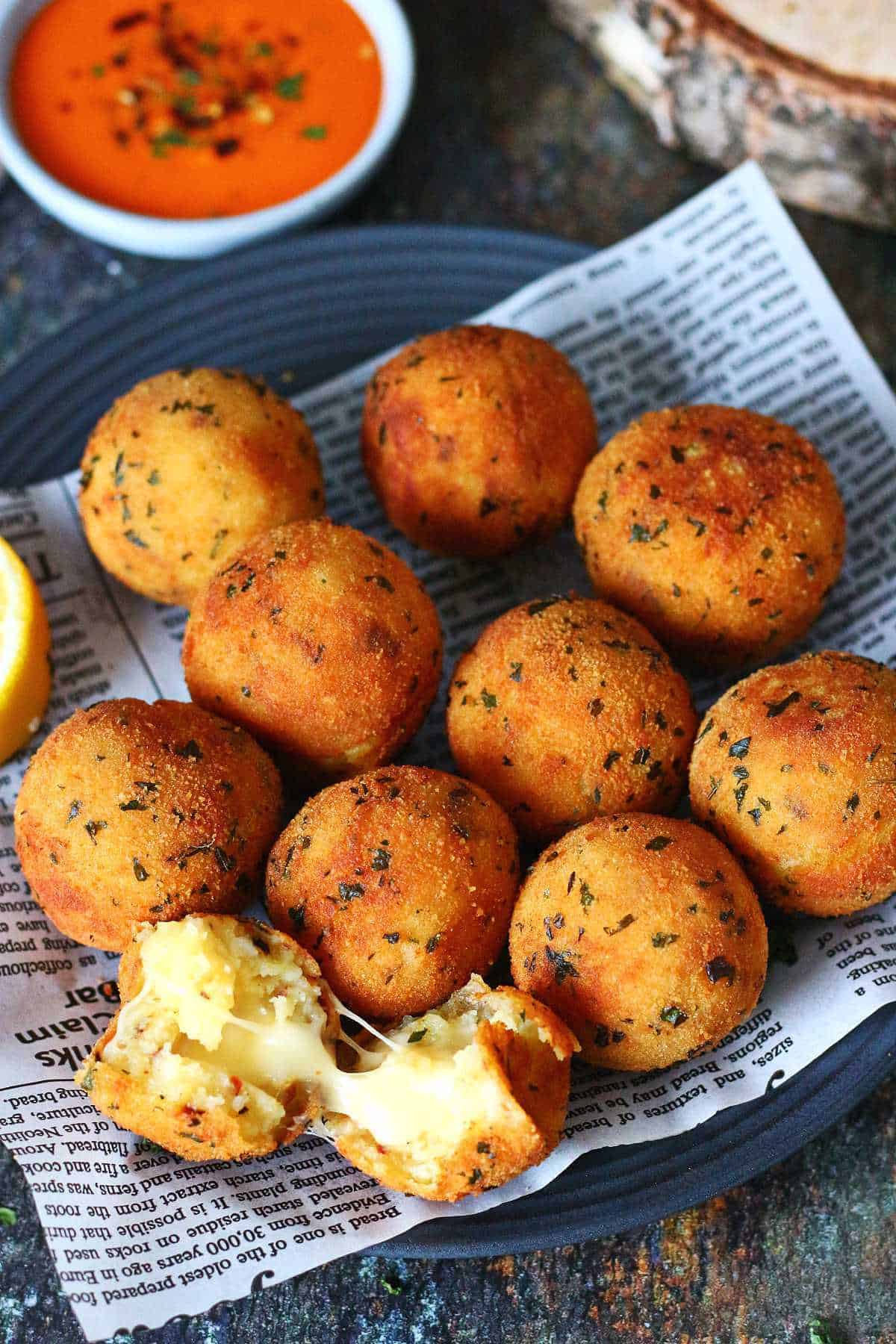 Plate of crispy potato and cheese balls on printed parchment paper with a small bowl of orange dipping sauce.