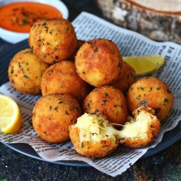 Stacked golden potato and cheese balls on a dark plate lined with printed paper, one ball split open showing melted cheese inside.