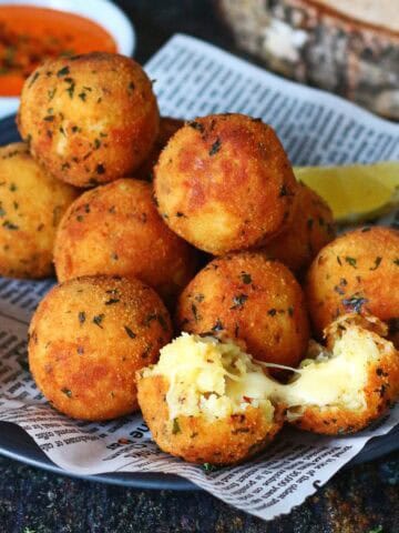 Stacked golden potato and cheese balls on a dark plate lined with printed paper, one ball split open showing melted cheese inside.
