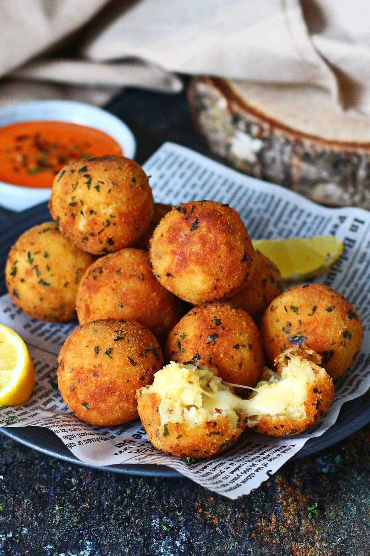Potato and cheese balls arranged on a plate with dipping sauce in the background and a lemon wedge on the side.