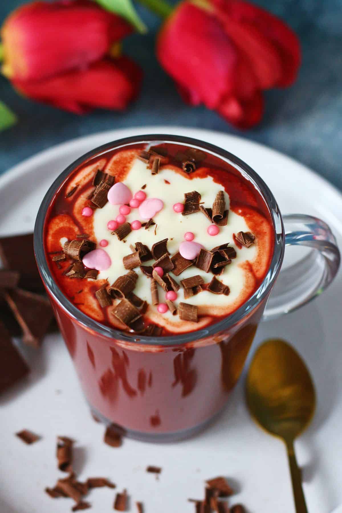 Glass mug filled with red velvet hot chocolate topped with cream cheese frosting, chocolate shavings, and pink sprinkles on a white plate.