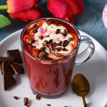 Glass mug of red velvet hot chocolate with cream cheese frosting, chocolate curls, and pink sprinkles, placed on a white plate with a gold spoon.