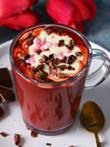 Glass mug of red velvet hot chocolate with cream cheese frosting, chocolate curls, and pink sprinkles, placed on a white plate with a gold spoon.