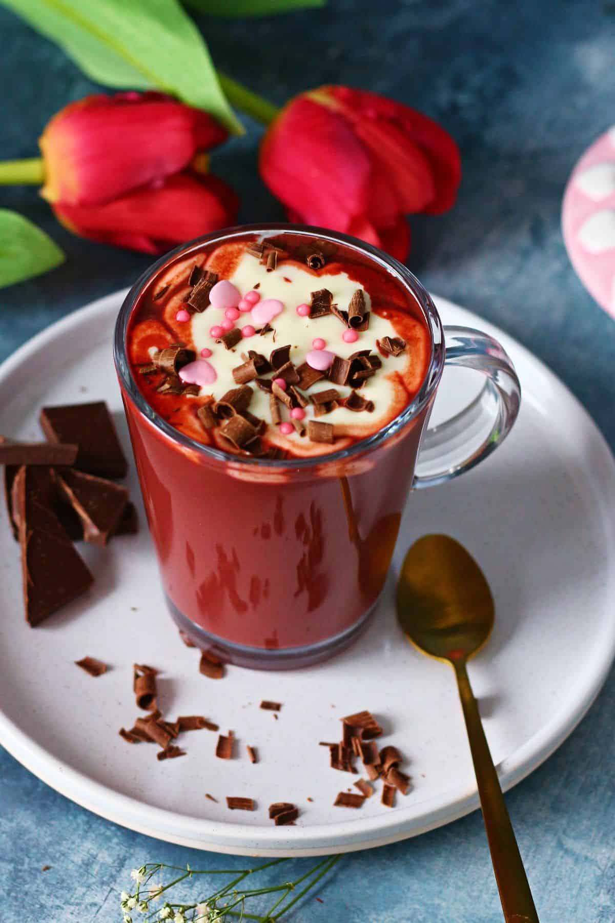 Glass mug of red velvet hot chocolate topped with cream cheese frosting and chocolate shavings, surrounded by red tulips .